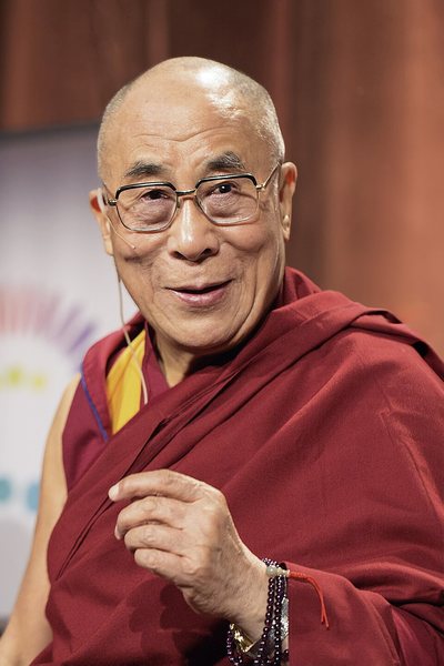 The 14th Dalai Lama in maroon monastic robes and glasses, wearing a headset microphone, smiling and gesturing with one hand