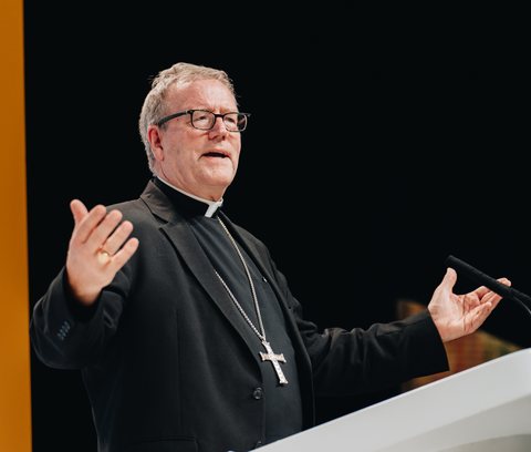 Bishop Robert Barron in a black clerical suit and Roman collar, wearing glasses and a large silver pectoral cross, gesturing with both hands raised while speaking from a lectern against a dark background