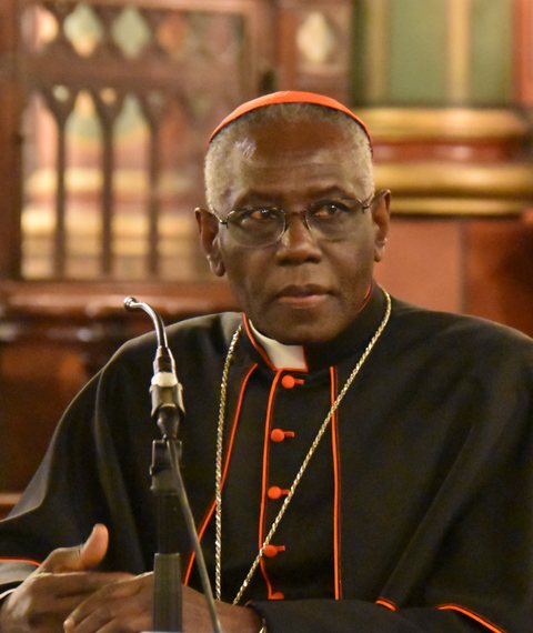 Cardinal Robert Sarah, an elderly Black man in glasses, seated at a microphone in a wood-panelled hall, wearing a black cassock with red piping and buttons, a red zucchetto, and a gold chain